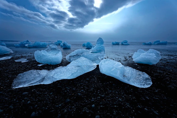 Icebergs glisten on a dark beach beneath cloudy skies.